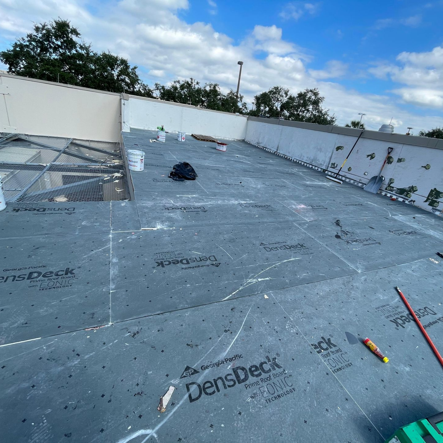 Flat commercial roof with dark, textured membrane; tools and supplies scattered. Cloudy sky overhead.