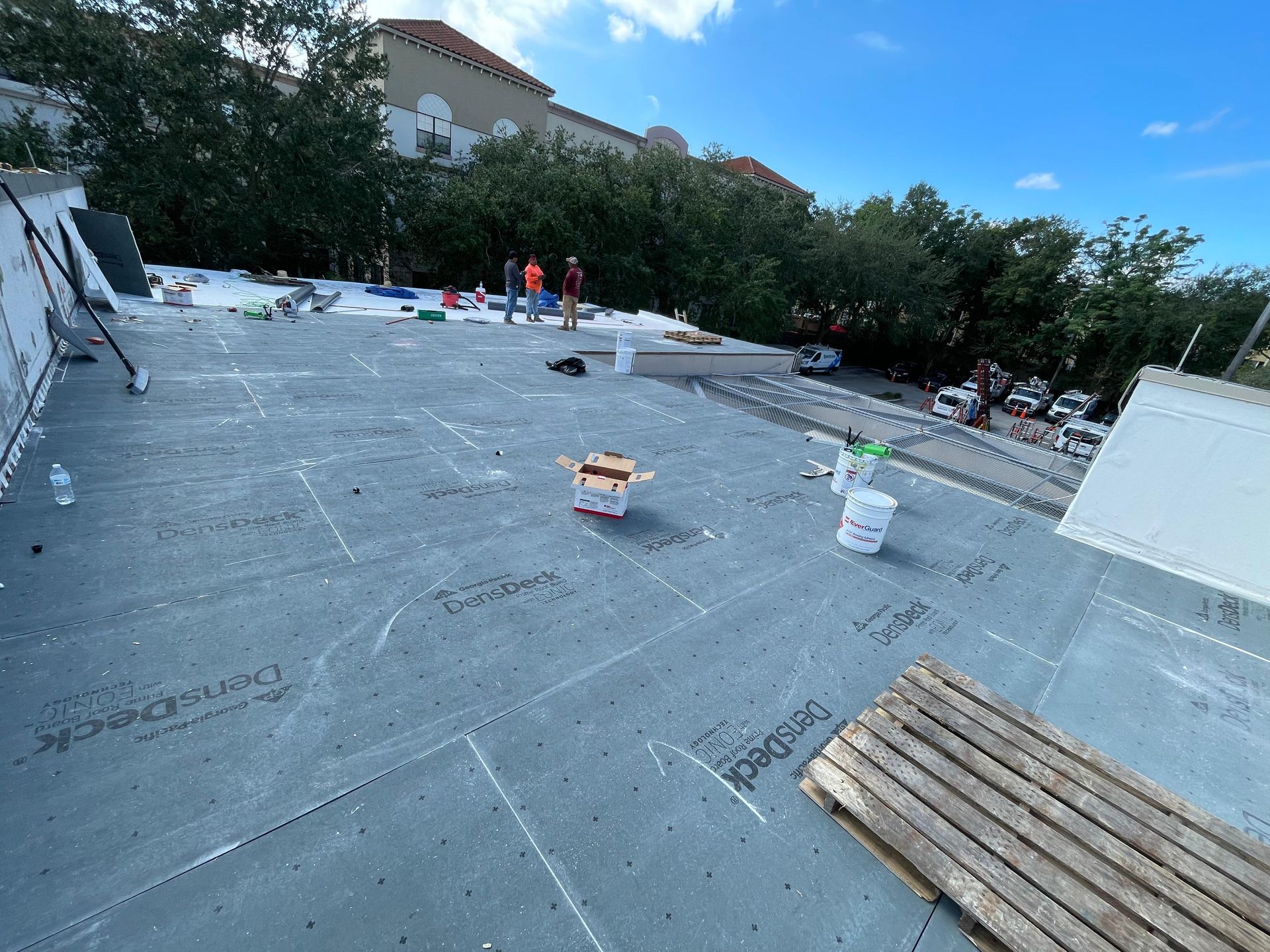 Rooftop construction: workers on a flat roof with gray membrane, buckets, and materials. Bright sky and trees.