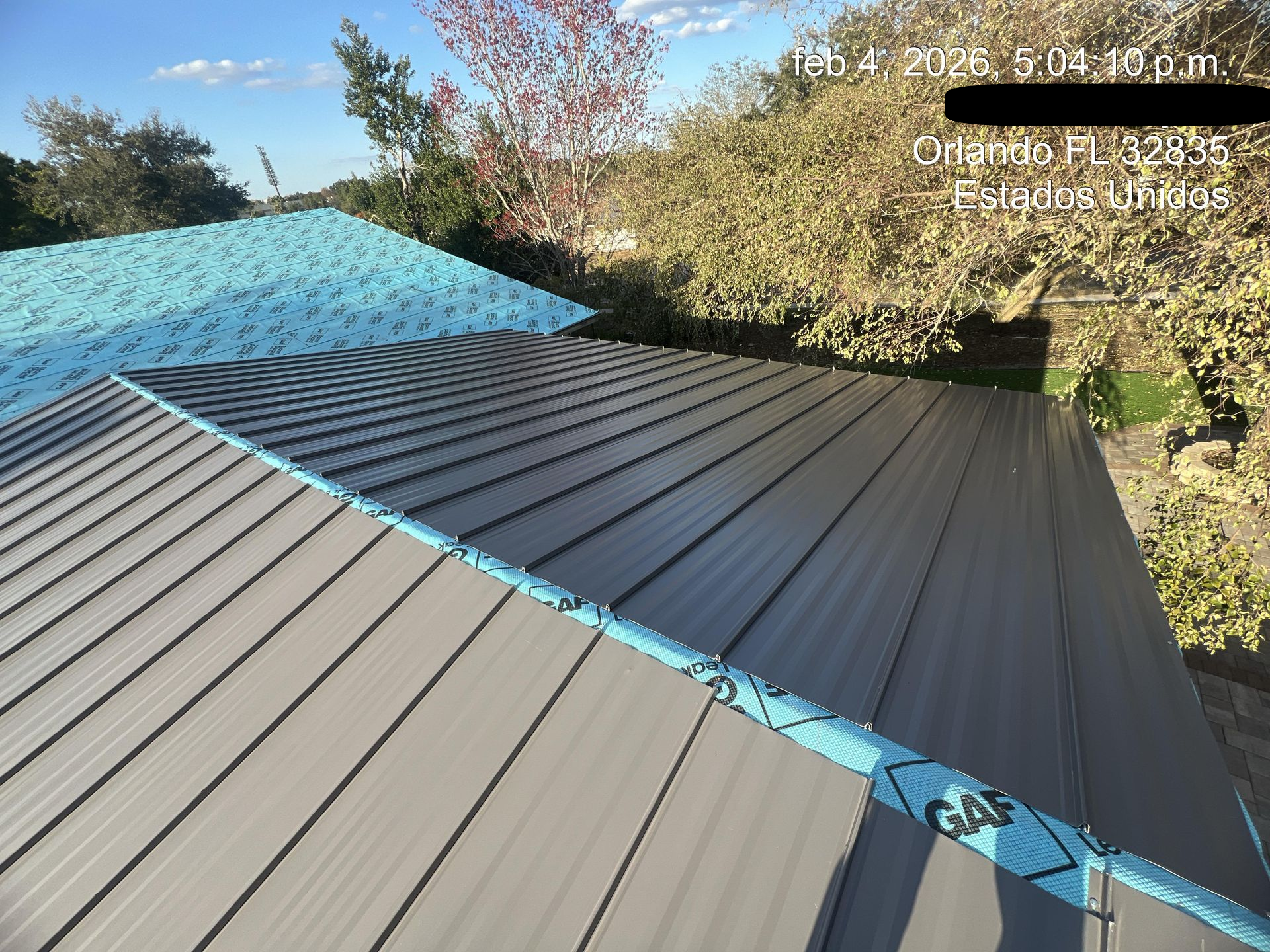 Blue and gray metal roof panels under construction beside trees and a garden path