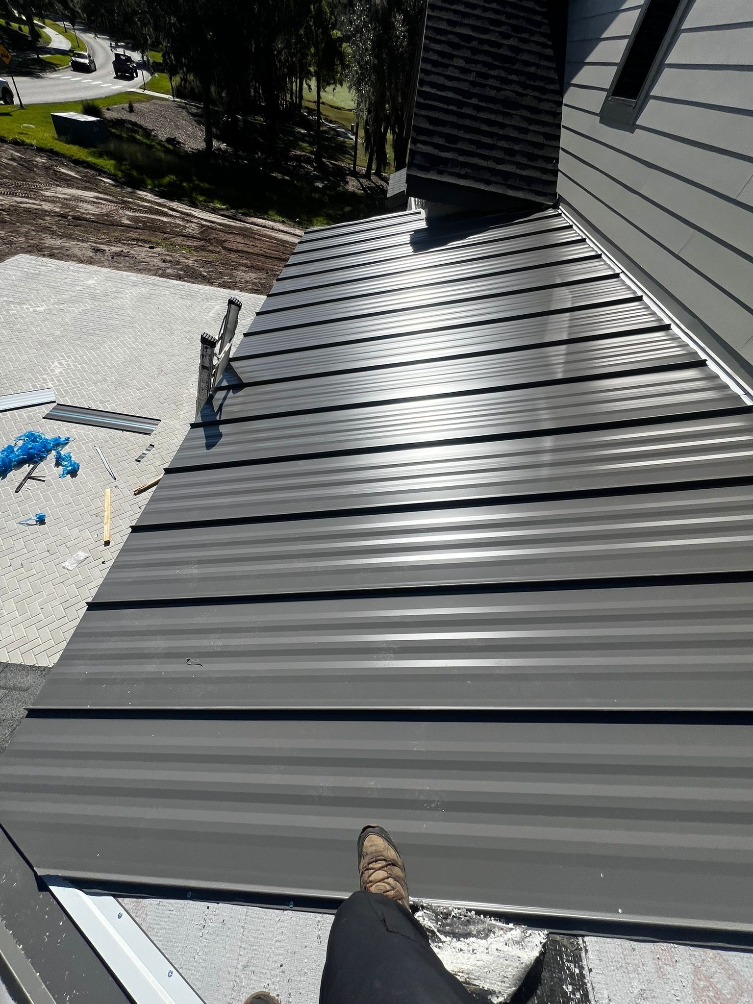 Person's legs and foot on a gray metal roof, possibly for a home. Sun reflecting on the roof.
