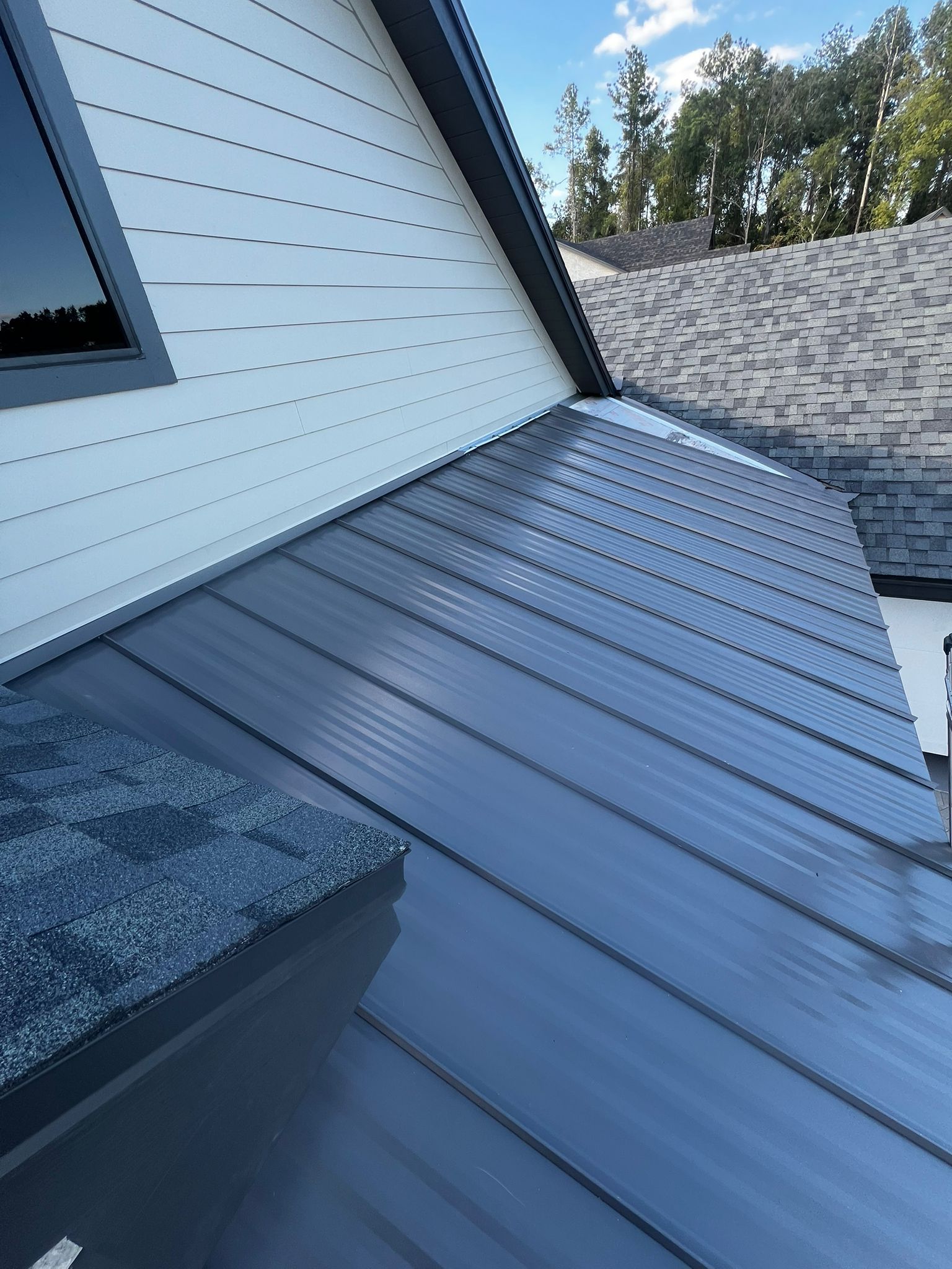 Close-up of a house roof with dark gray metal panels, white siding, and a small window.