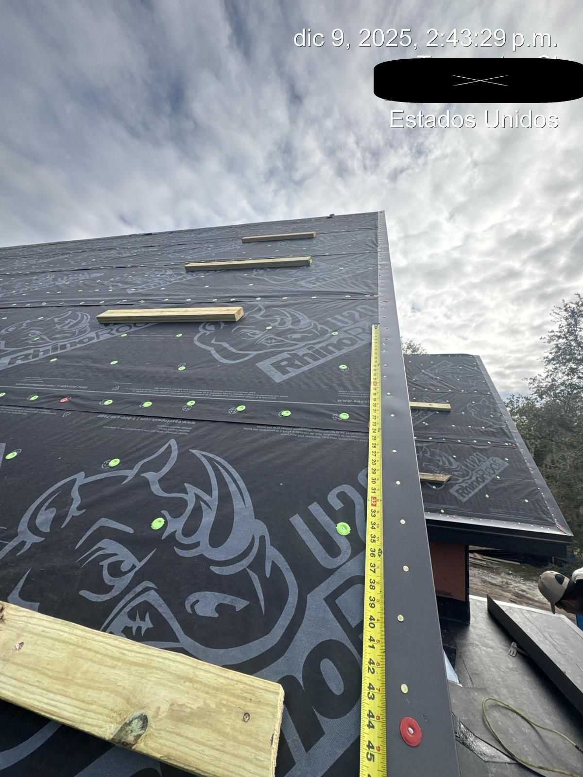 Rooftop under construction. Black underlayment, wooden supports, and yellow measuring tape. Cloudy sky overhead.