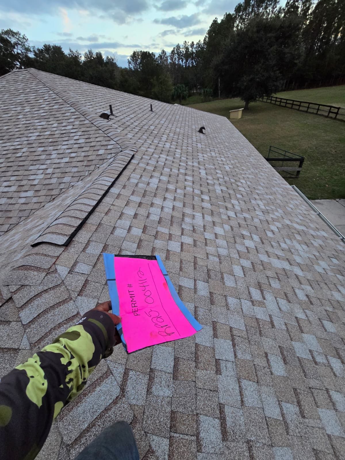 Person on a roof holds a pink clipboard, view of shingles, sky, and grass with a fence.