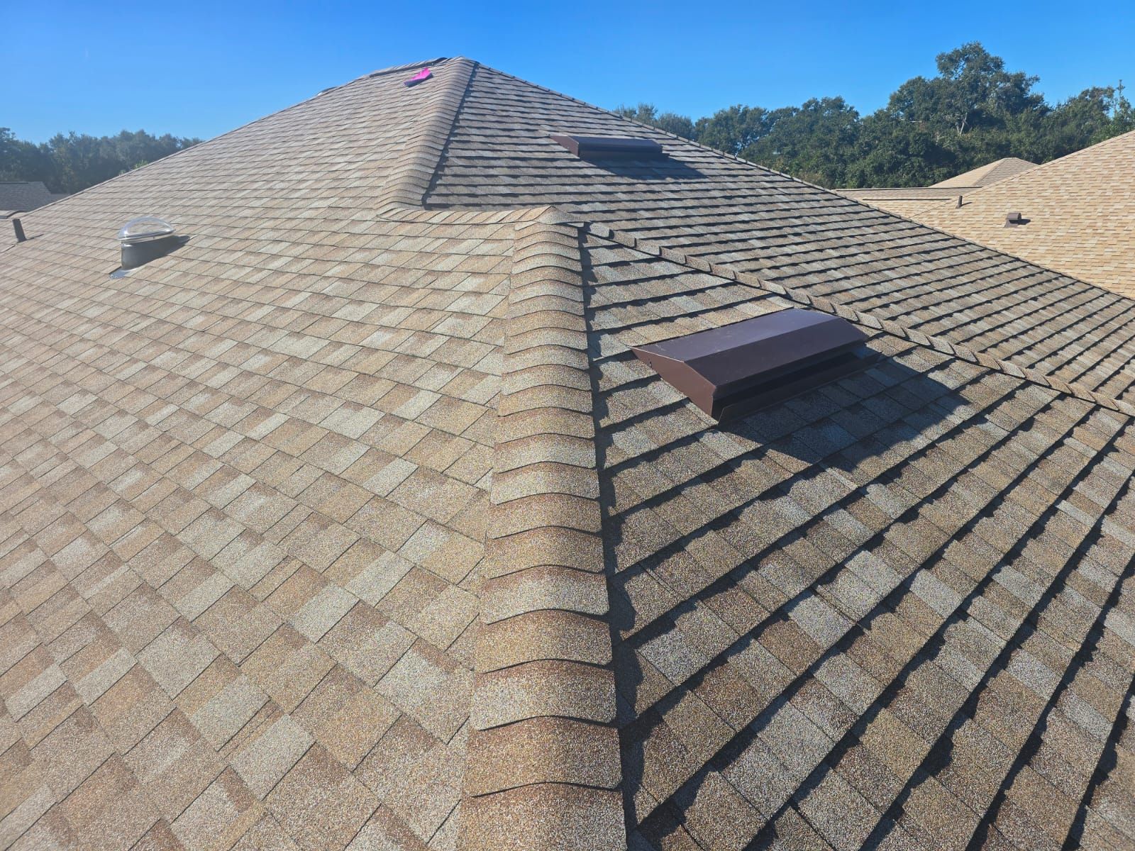 Brown shingled roof with a skylight and vent against a clear blue sky.