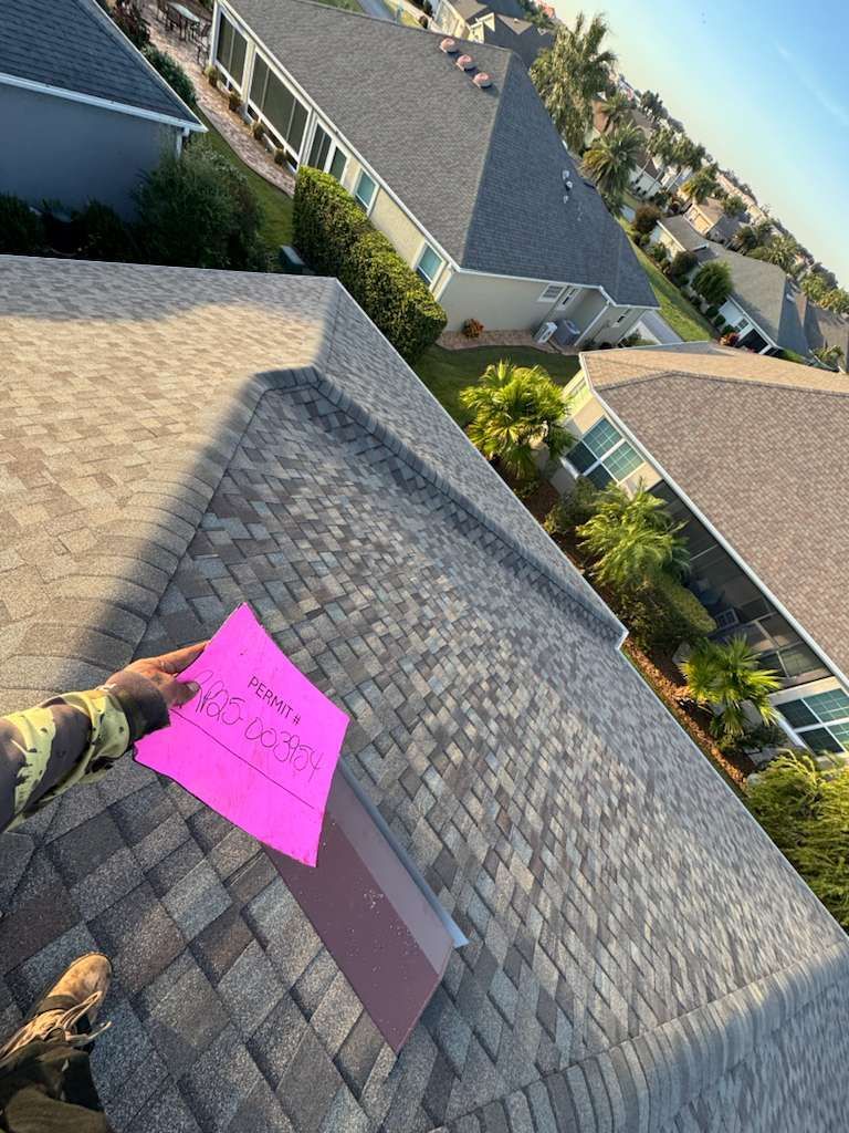 Person holding pink paper on a shingled roof, overview of suburban houses under a blue sky.