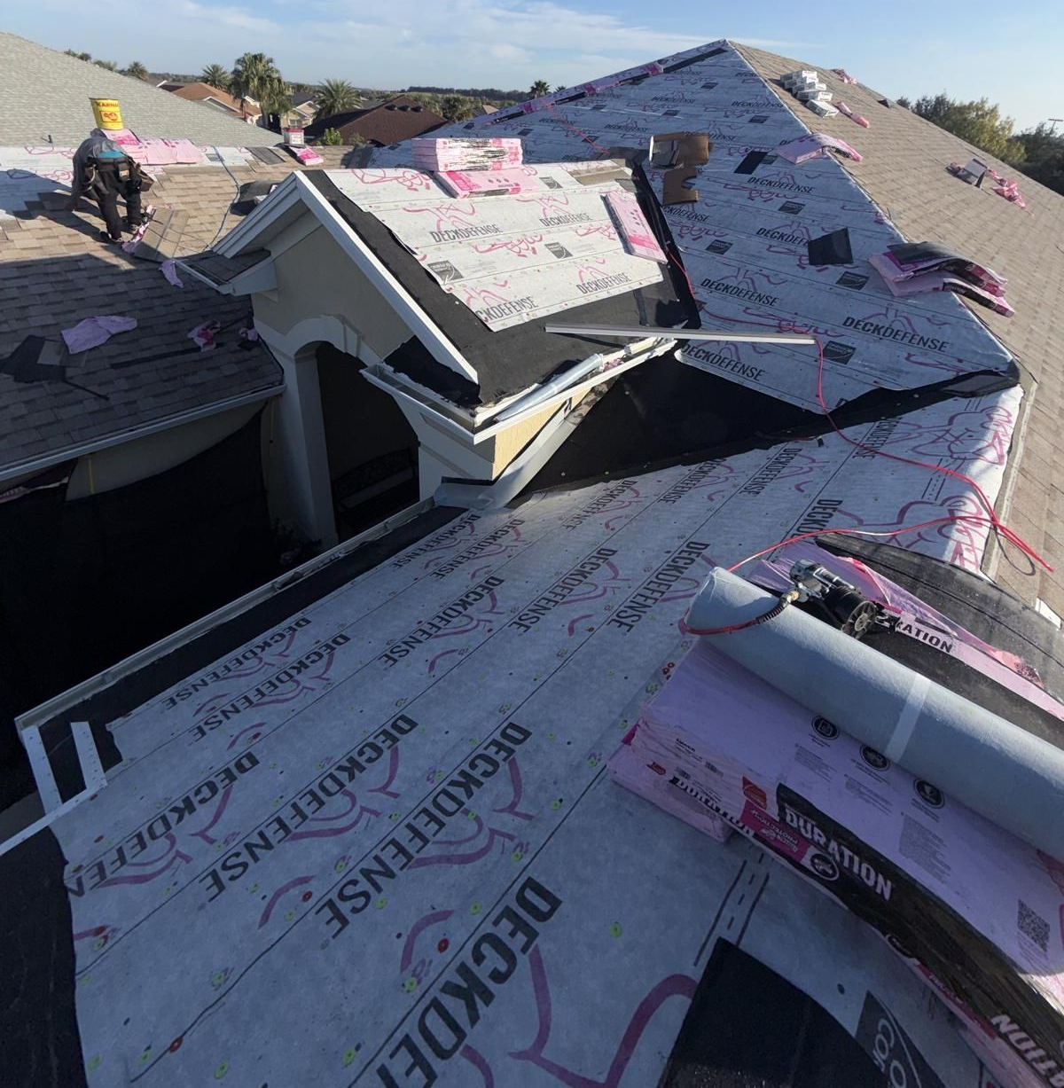 Roof being repaired; roofing materials visible; worker on roof; sunny day.