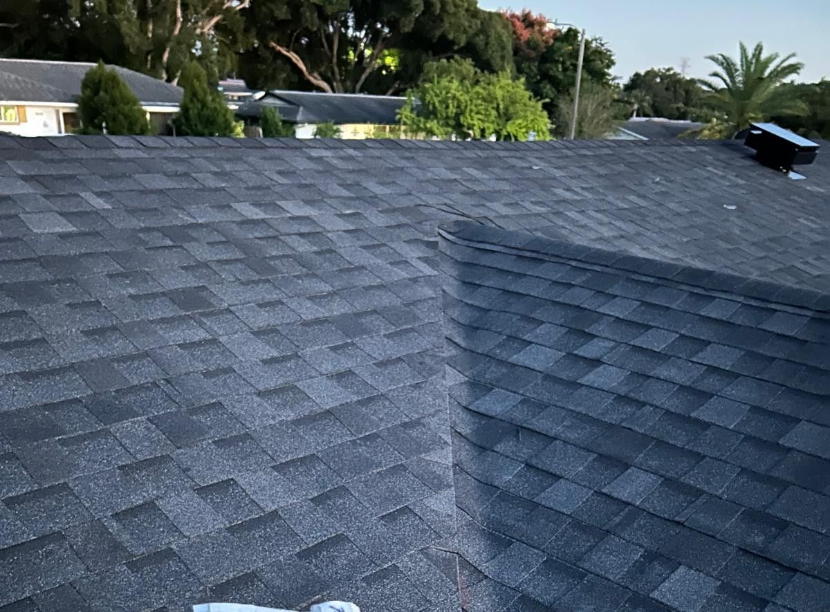 Dark gray asphalt shingle roof, angled view. Buildings and trees visible in background.