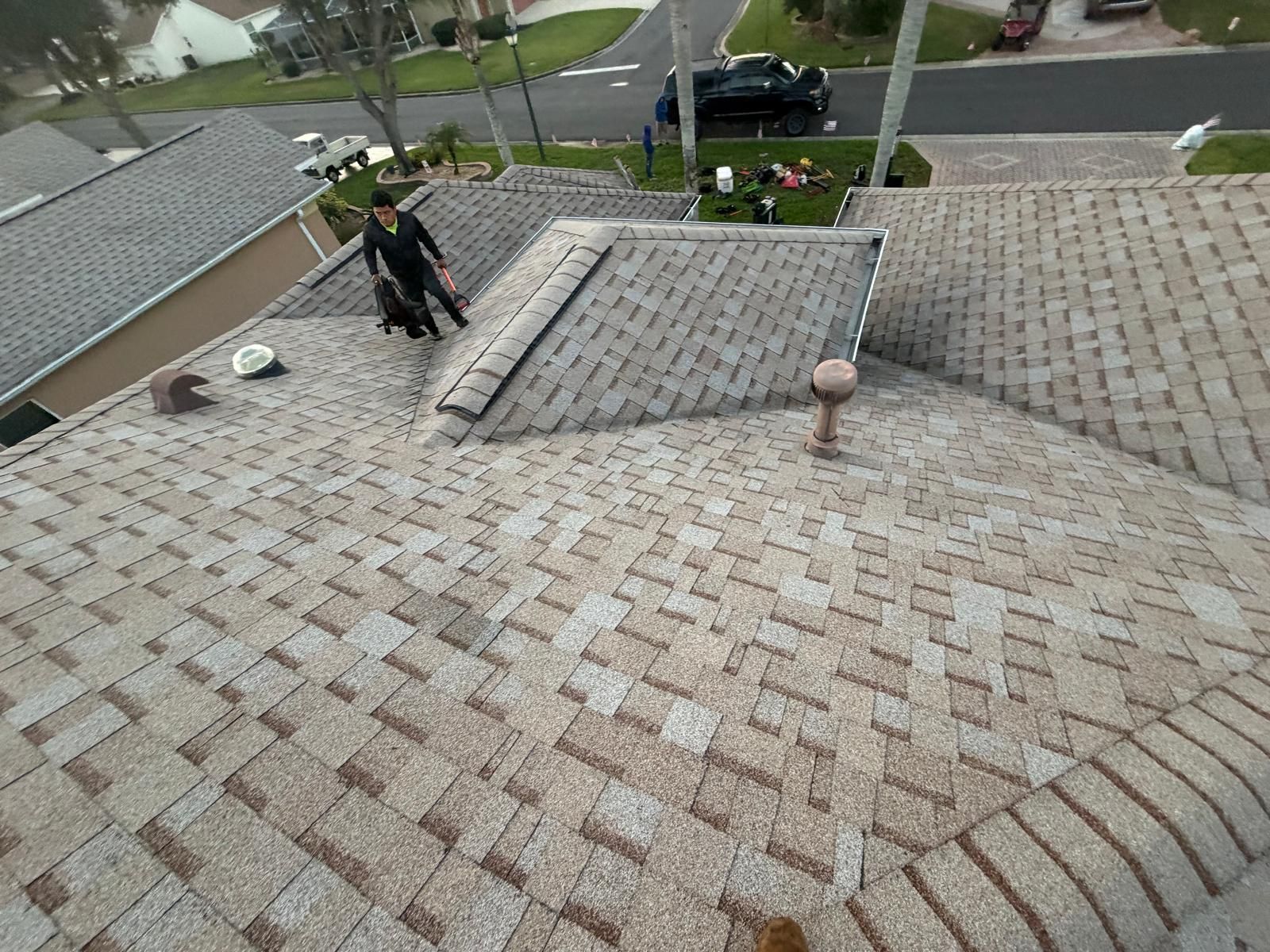Man on a roof, inspecting shingles. Overlooking neighborhood with parked cars and green lawns.
