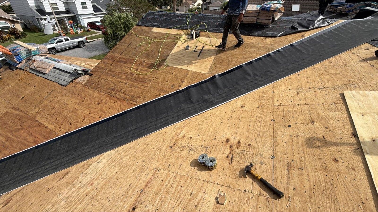Roofer installing roofing materials on a wooden roof. Black underlayment, hammer, and plywood are visible.