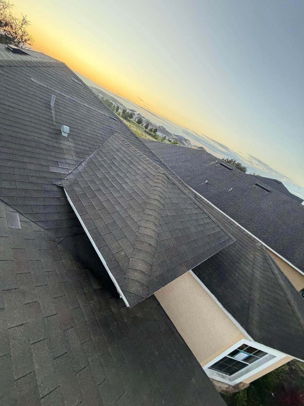 Black shingle roof with a chimney and skylight, viewed from above against a sunrise sky.