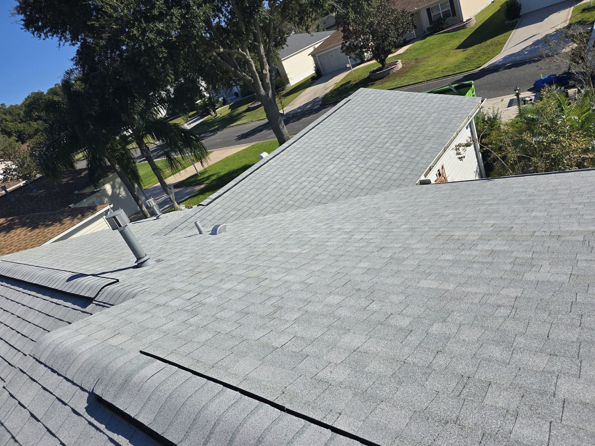 Gray asphalt shingle roof on a house, angled view, with trees and a sunny sky.