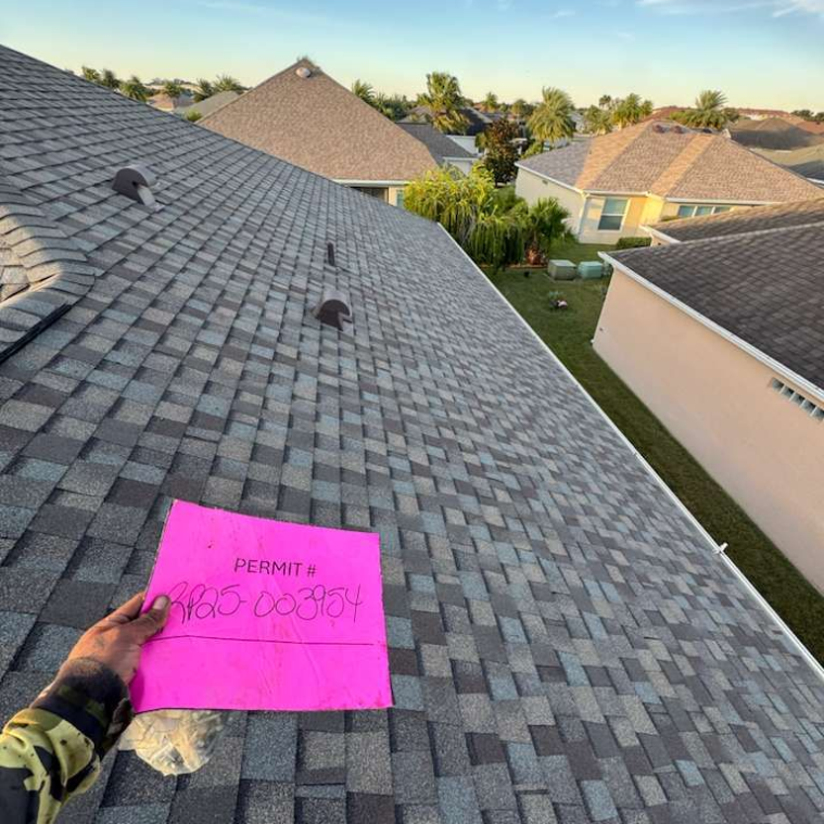 Roofer holding a pink permit on a newly shingled roof. Nearby are other houses and greenery.