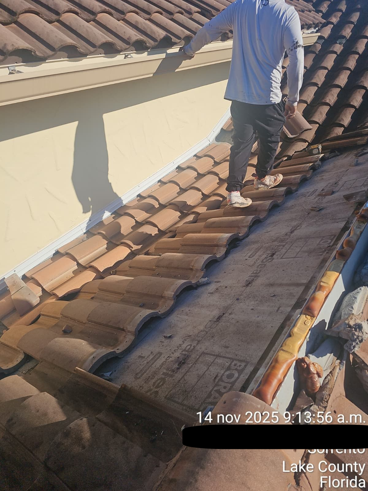 A person walks on a tiled roof. Light blue shirt, dark pants, and white shoes. Beige walls and shadows.