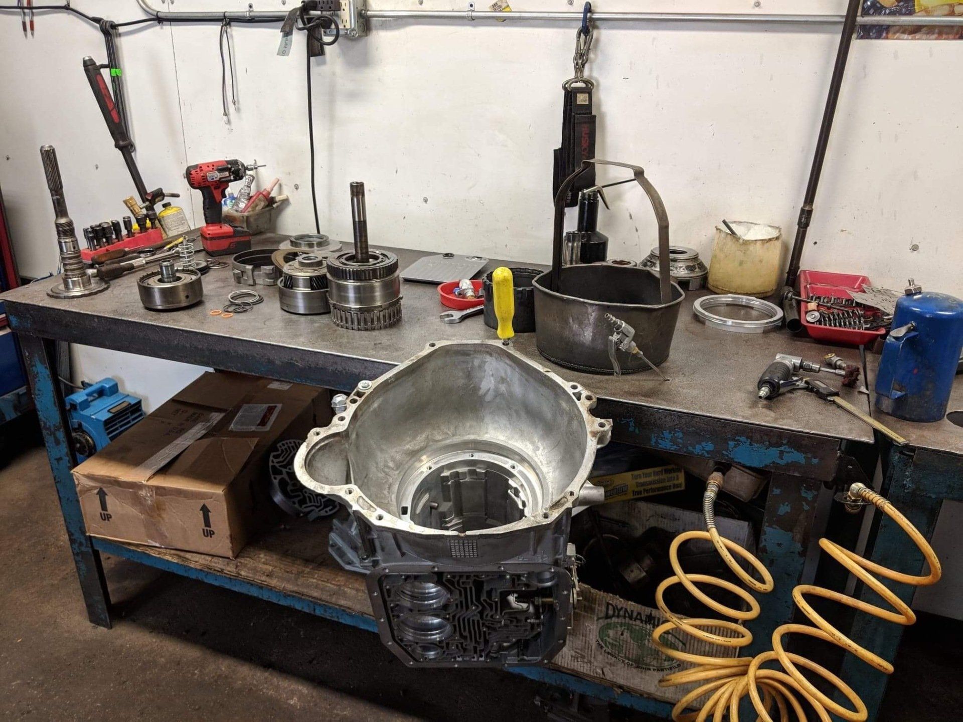 Cluttered workshop bench with metal parts, tools, a large silver basin, and coiled yellow air hoses