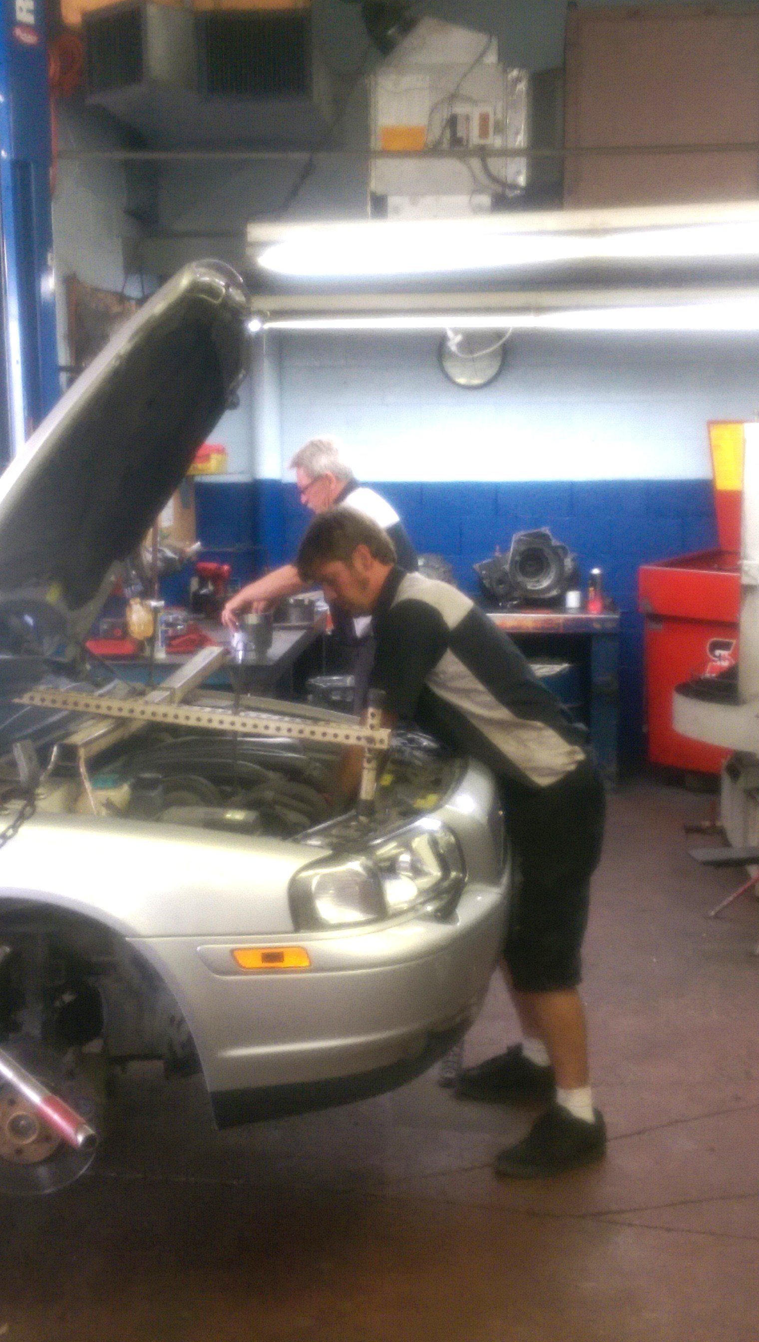 Person leaning over a silver car with hood open in an auto repair shop.