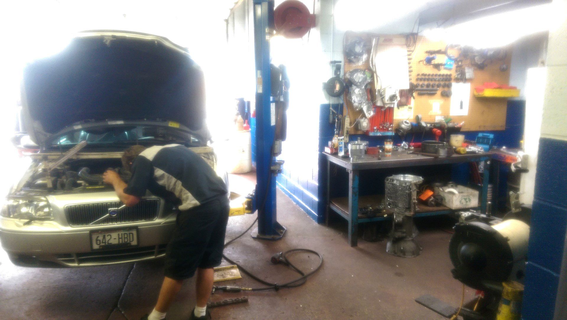 Car in a garage with hood open; person leaning over the engine beside tools and equipment.