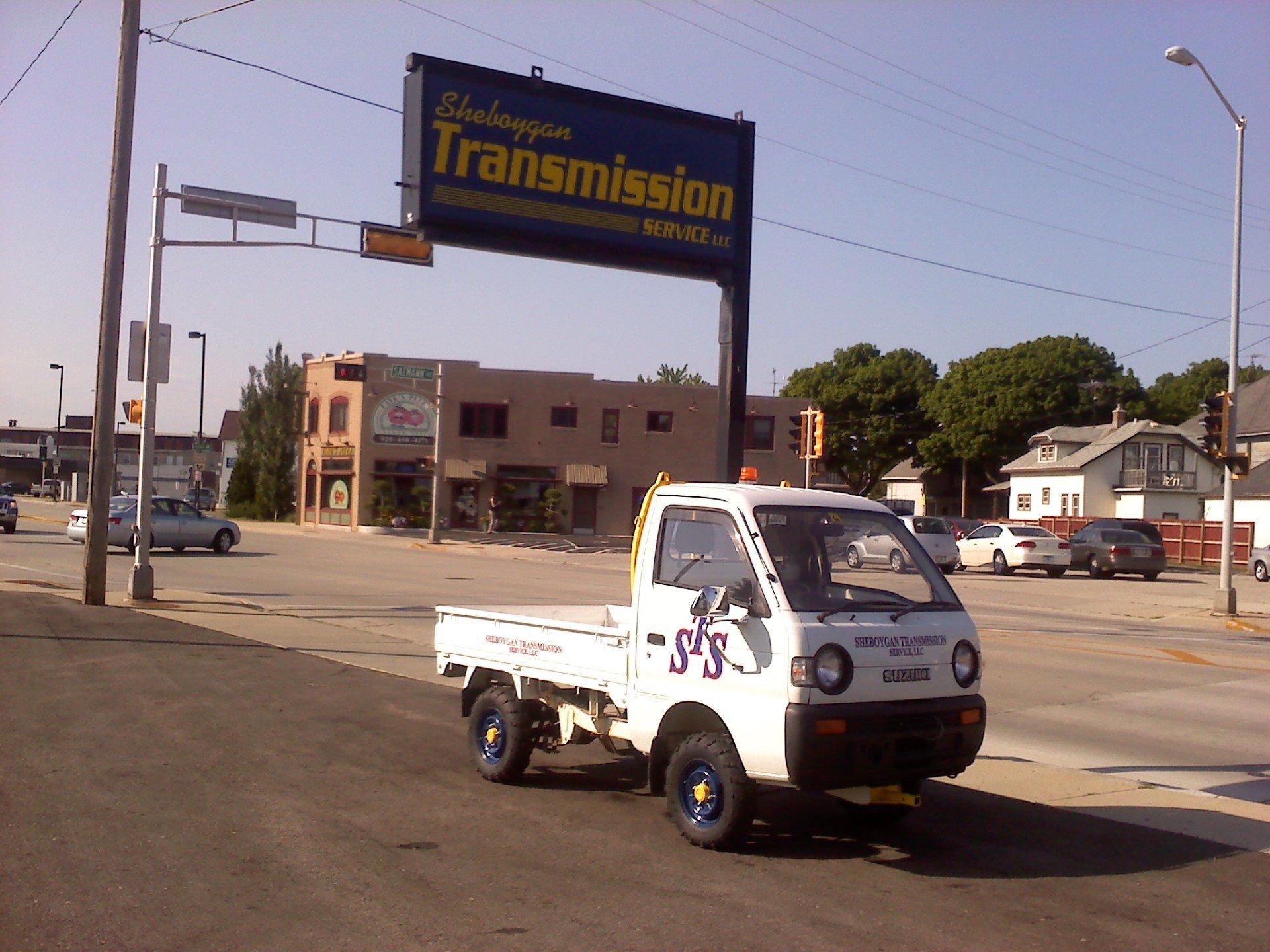 Small white pickup truck driving past a transmission shop sign at a street intersection.