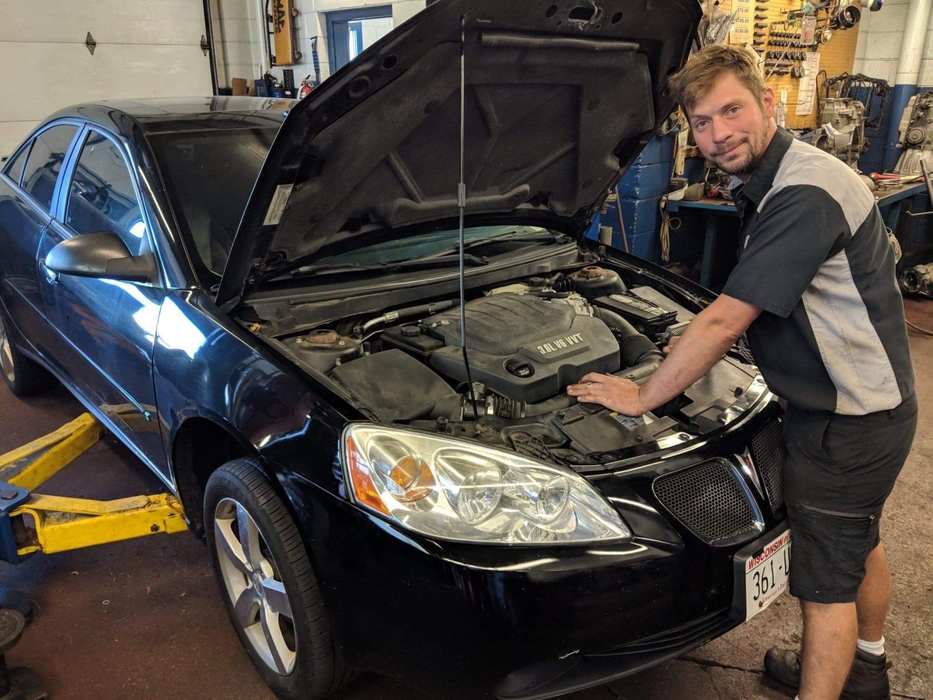 Man in a garage leaning over the open hood of a black sedan on a lift