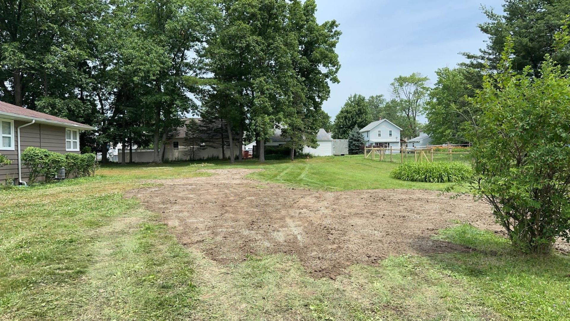 Bare yard with a dirt patch, green trees, and houses in the background on a sunny day