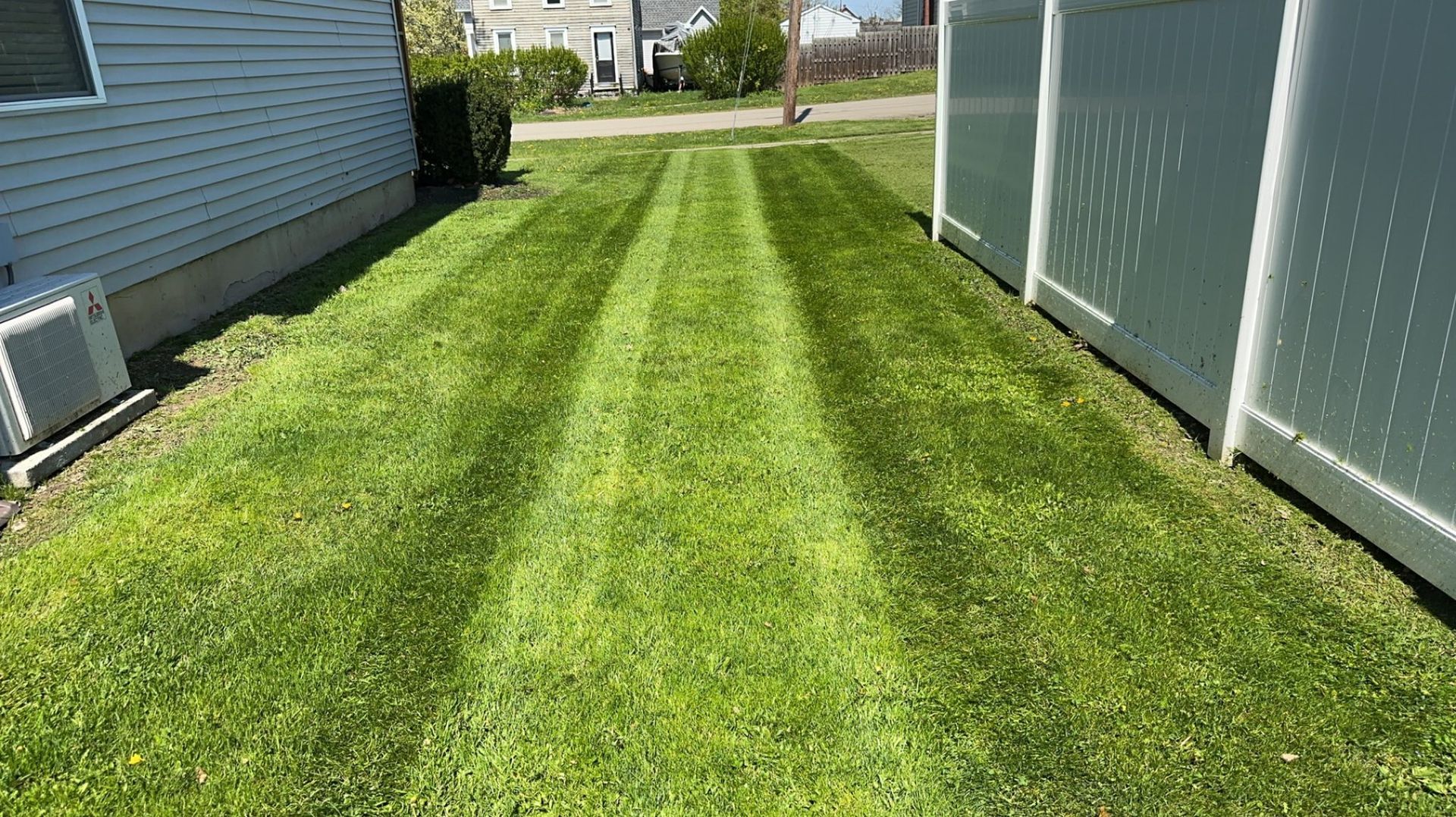 A narrow green lawn featuring fresh, parallel mowing stripes between the side of a house and a white privacy fence.