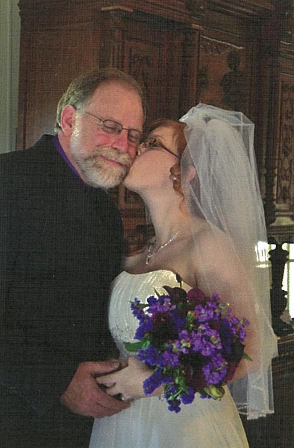 A bride is kissing her father on the cheek while holding a bouquet of purple flowers.