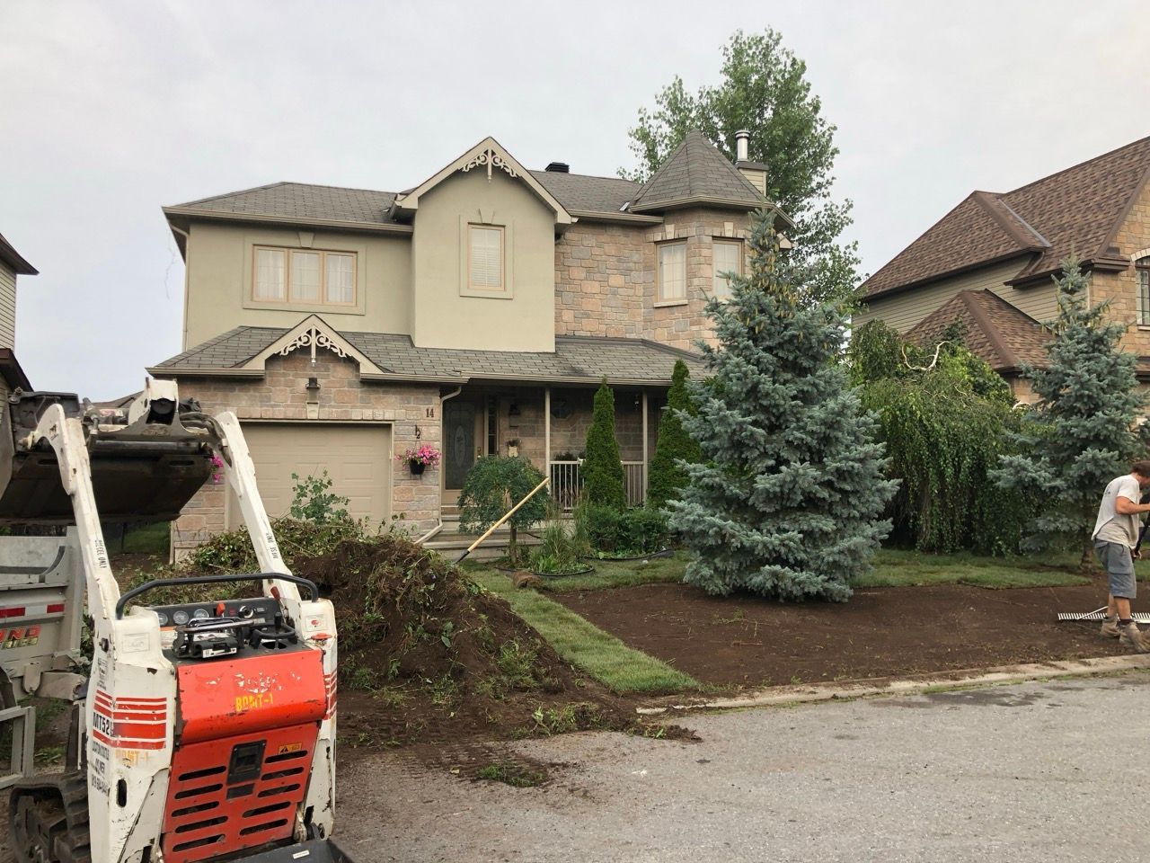 Aménagement paysager en cours dans une maison à deux étages; chargeuse Bobcat, gazon fraîchement posé, arbres et un ouvrier.
