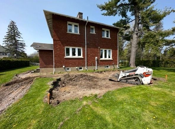 Une maison en briques avec une petite excavatrice sur l'herbe ; le sol est creusé pour une éventuelle construction.