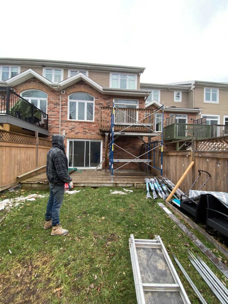 Un homme dans son jardin regarde un échafaudage sur une terrasse. Des maisons de ville en briques en arrière-plan, sous un ciel couvert.