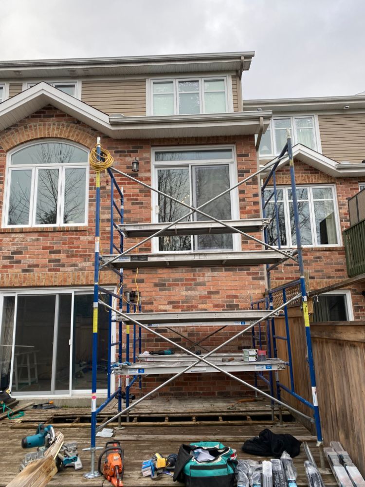 Échafaudage installé contre une maison en briques avec fenêtres, sur une terrasse en bois avec des outils.