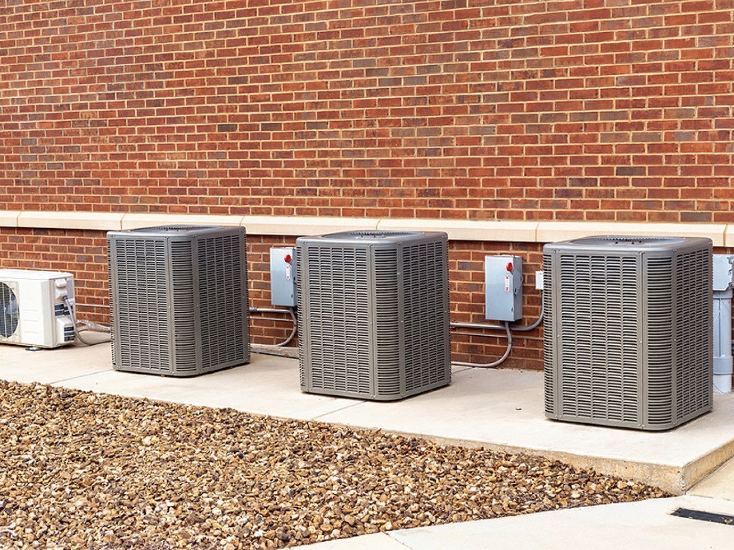 Three gray HVAC units sitting on a concrete pad against a brick wall.