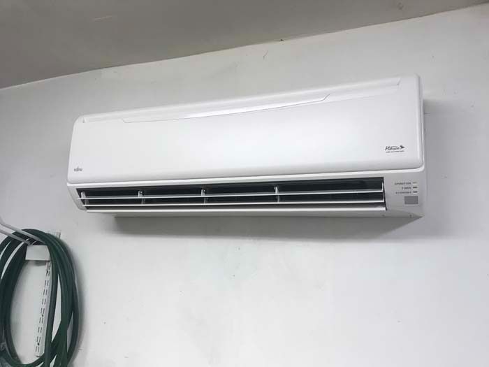 A white split-system air conditioner mounted on a plain white wall next to a coiled garden hose.