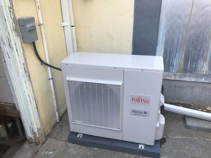A white Fujitsu outdoor HVAC heat pump unit installed on a concrete base next to a wall with an electrical disconnect box.