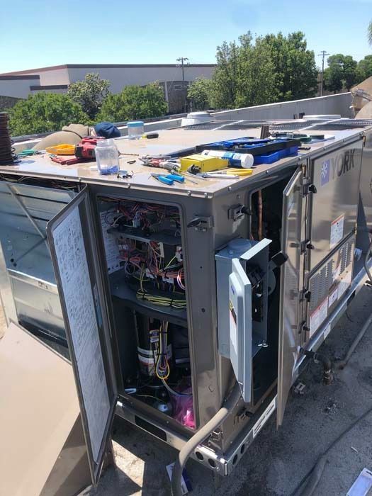 A rooftop HVAC unit with an open electrical panel and tools scattered on top under a clear blue sky.