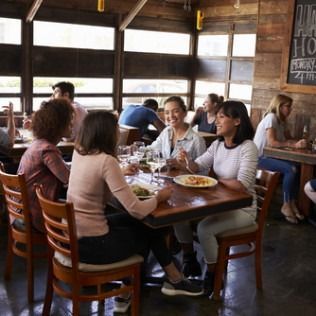 A group of people dining and chatting at a wooden table in a brightly lit, rustic restaurant.