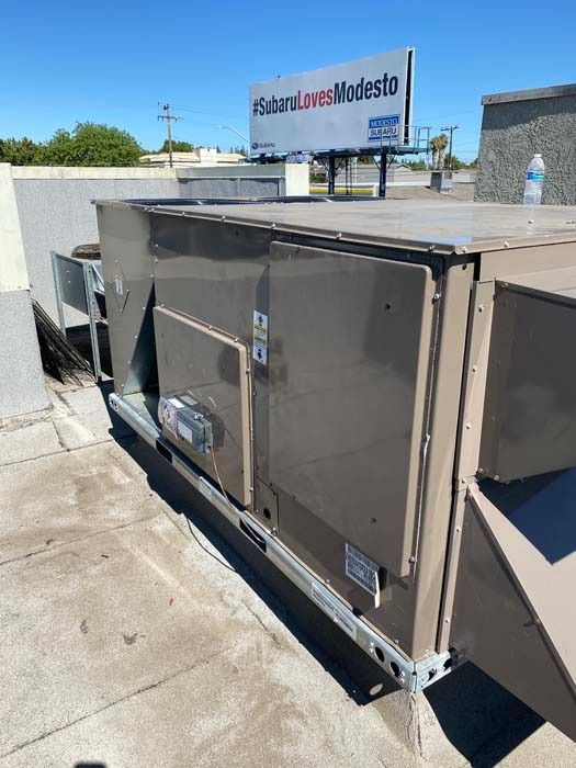 A tan industrial HVAC unit sits on a flat rooftop under a clear blue sky, with a billboard visible in the background.