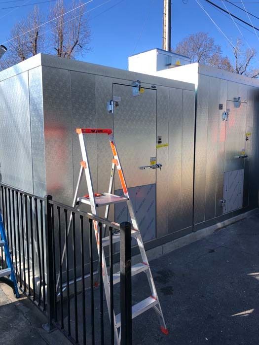 A metal outdoor utility enclosure with a ladder positioned in front of it under a clear blue sky.