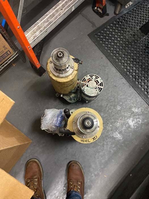 Two industrial pump motors sitting on a gray floor with a person’s brown work boots visible at the bottom.