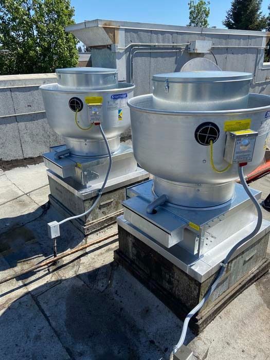 Two industrial exhaust fans installed on a flat, gravel-covered roof under a clear blue sky.