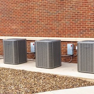 Three grey outdoor HVAC condenser units sit on a concrete pad against a red brick building wall.
