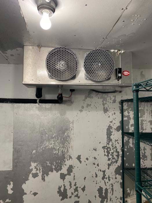 A walk-in cooler evaporator unit with two circular fans mounted on a wall with peeling gray paint.