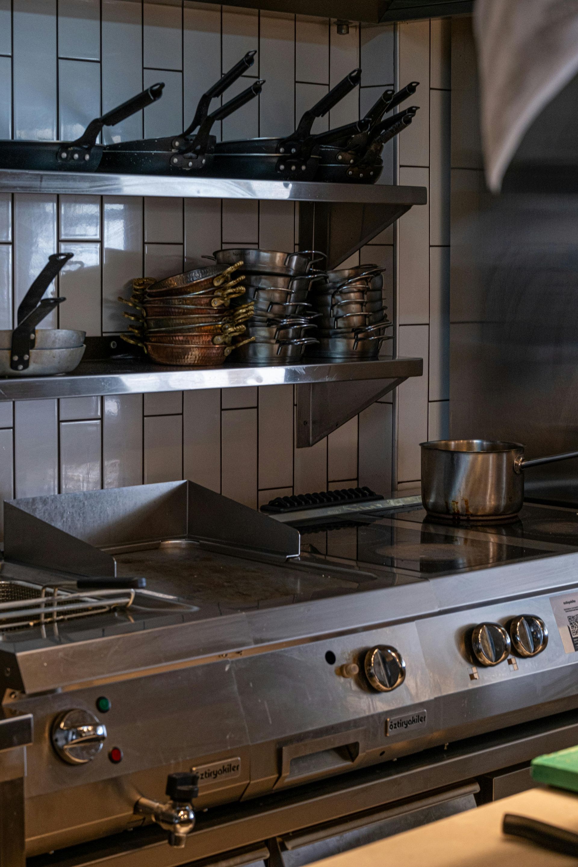 A professional stainless steel kitchen range with stacked cookware and pans on shelves against a tiled wall.