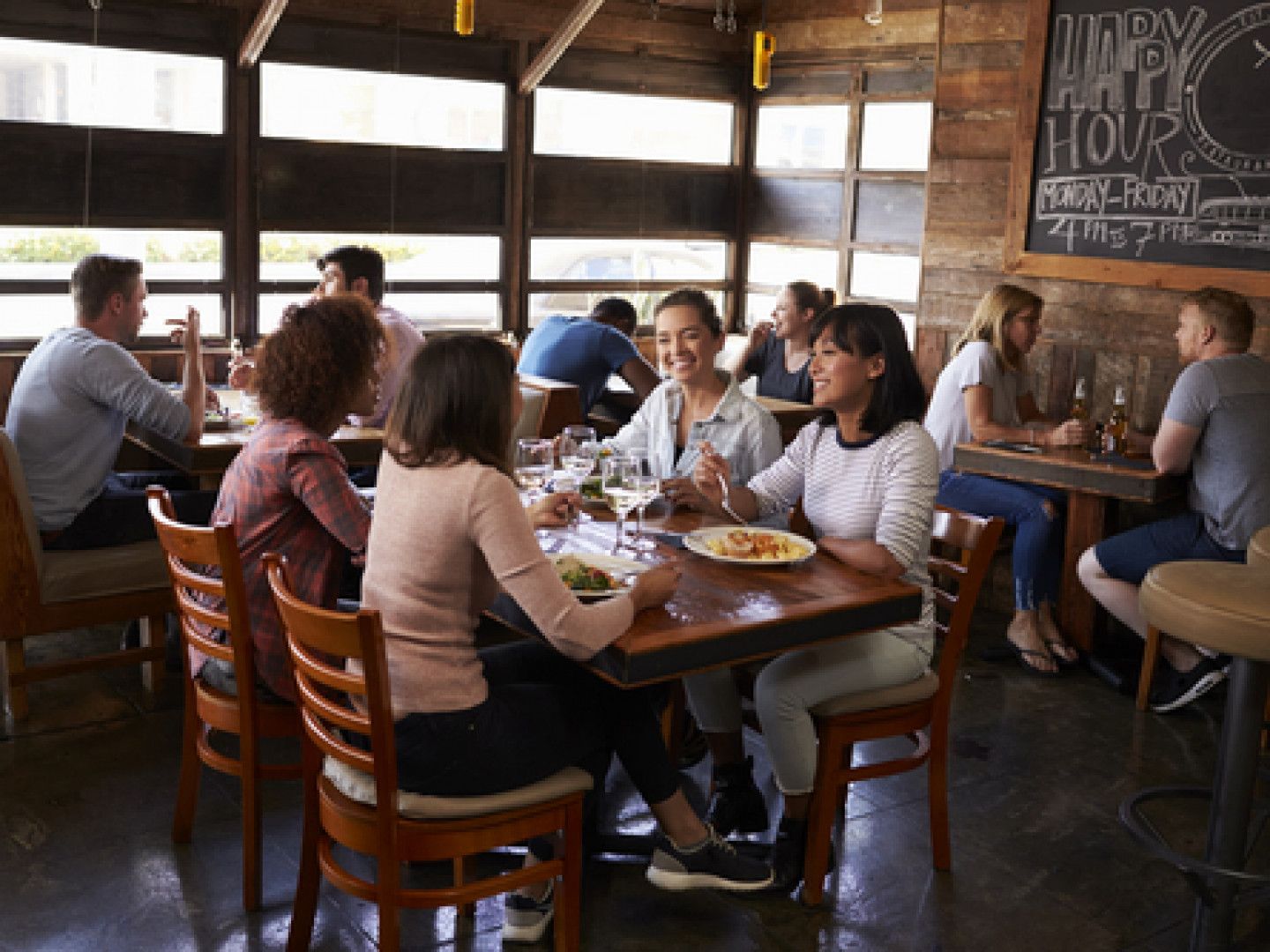 A group of people dining and chatting at wooden tables in a rustic, casual restaurant with a happy hour chalkboard.