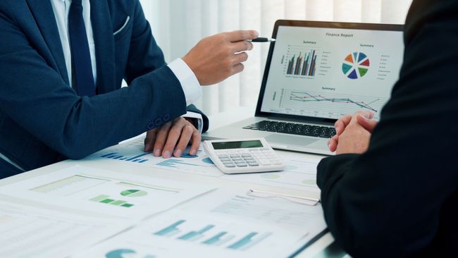 Two people reviewing financial charts on a laptop at a desk, with printed reports spread out.
