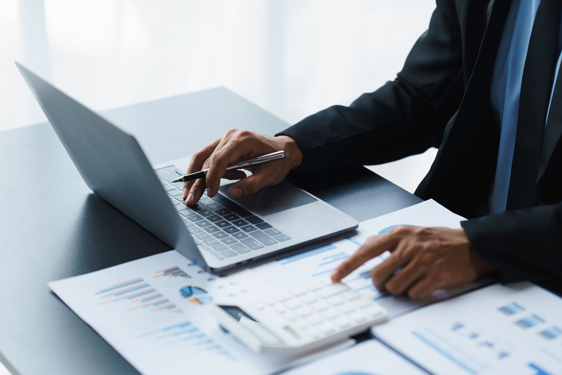 Person typing on a laptop beside printed charts and documents in a bright office setting