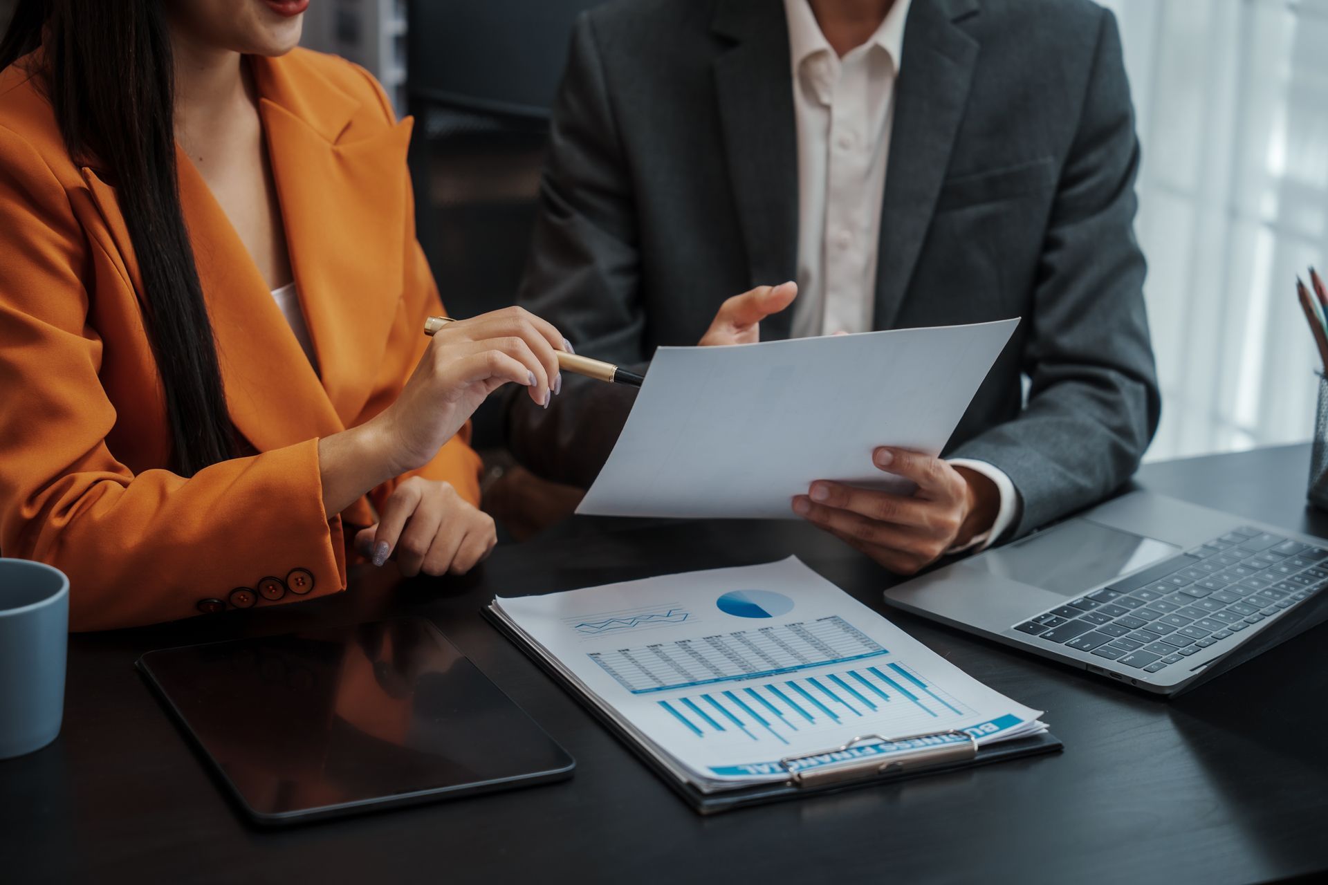 Two people in business attire reviewing charts and documents at a conference table.