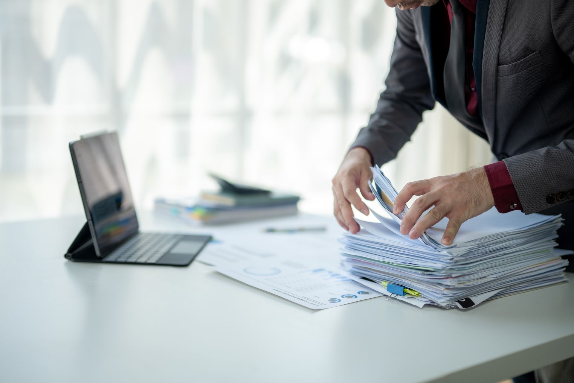 Person sorting a stack of papers beside a laptop on a bright office desk
