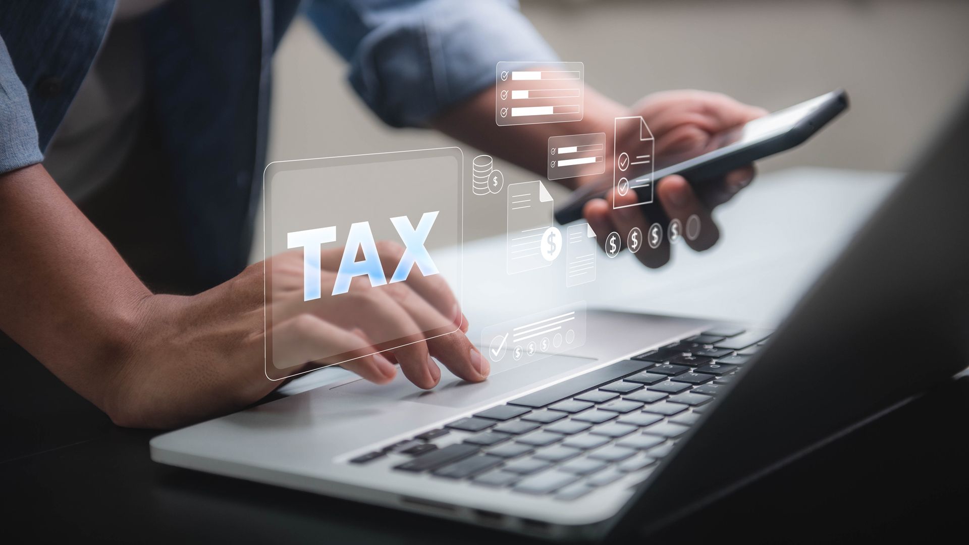 Hands typing on a laptop beside a smartphone with a glowing TAX overlay, suggesting online tax filing.