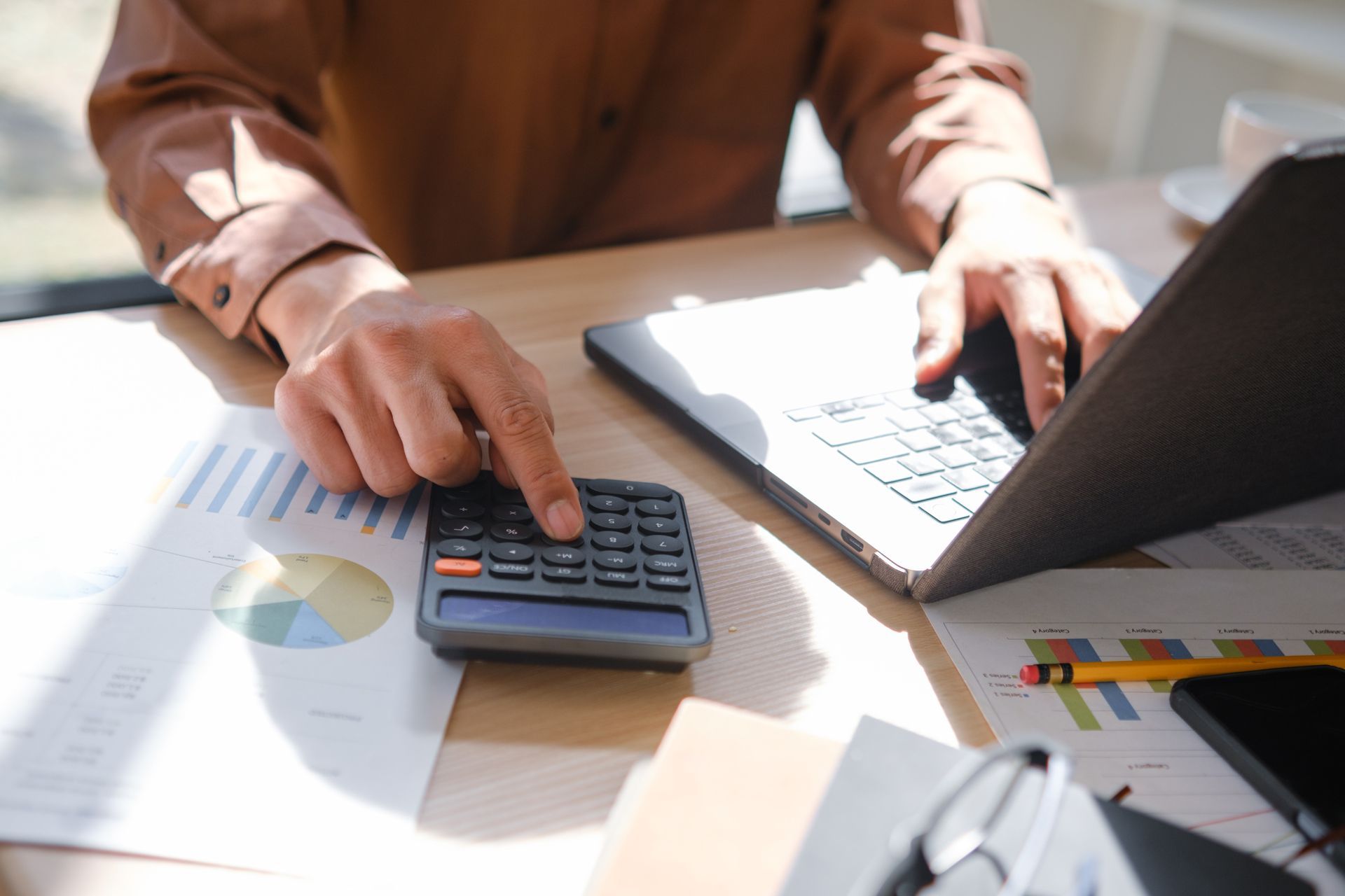 Hands using a calculator beside a laptop on a desk with spreadsheets and charts