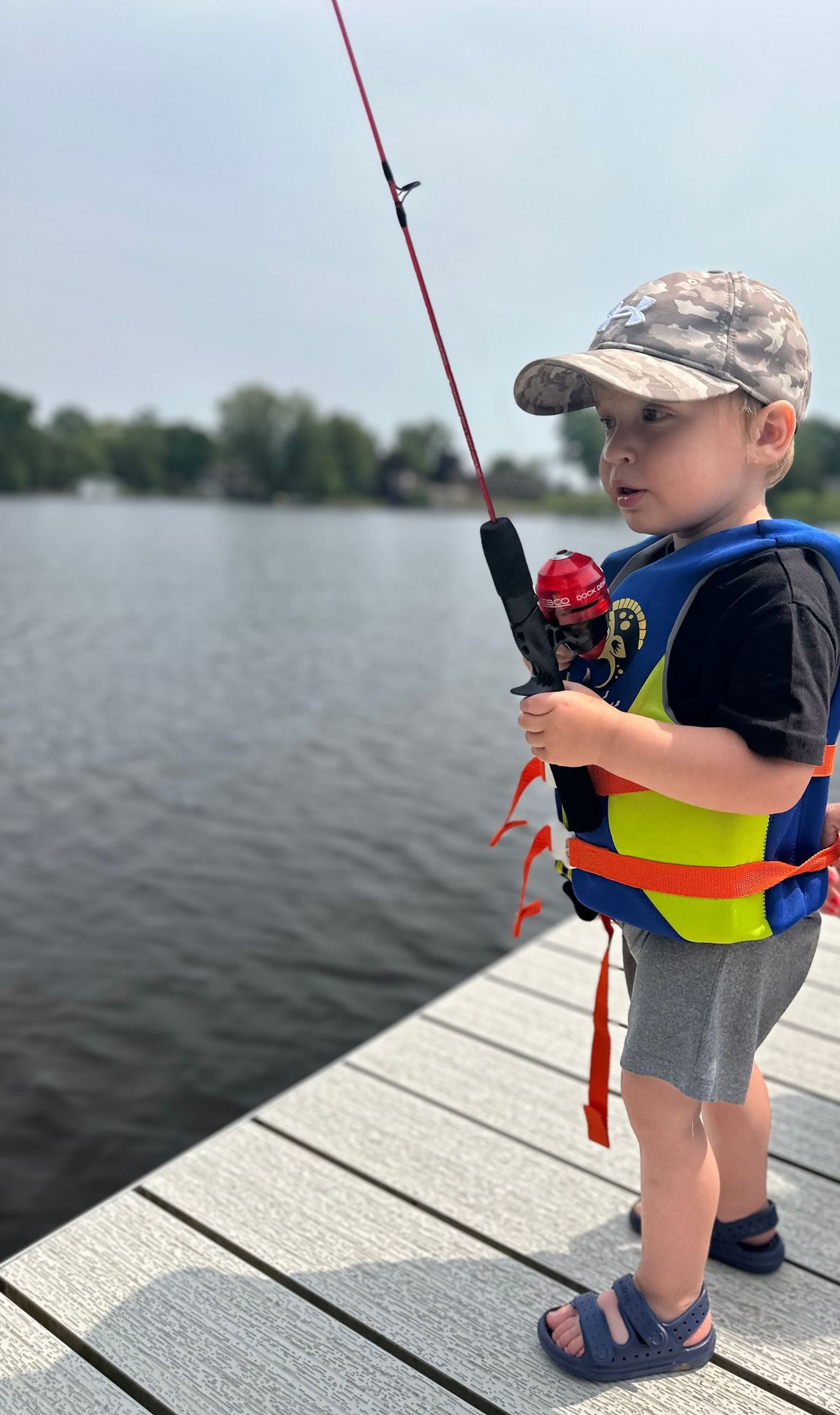 Child fishing on a dock, wearing a camo cap and bright life vest beside a calm lake.