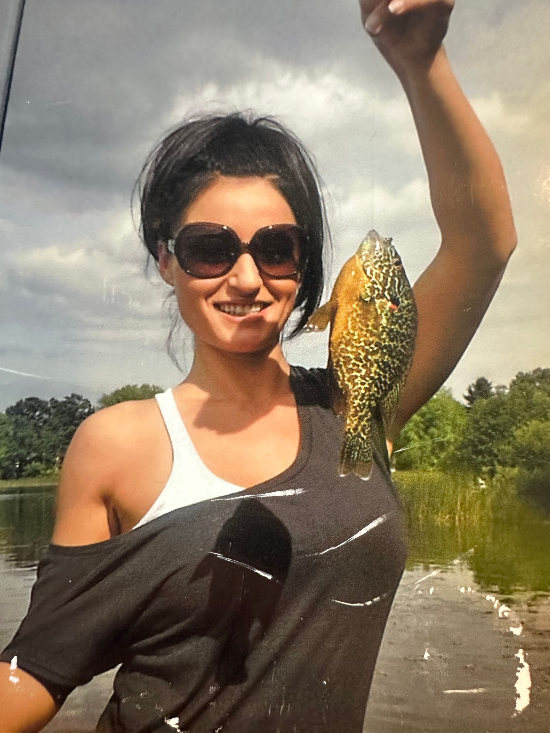 Person holding up a fish while smiling outdoors near a river, wearing sunglasses and a white tank top.